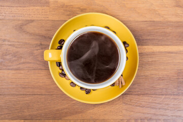 coffee composition, aerial shot of a yellow ceramic cup full of single coffee, on the coffee plate there are coffee beans and a cinnamon stick, the background surface is wooden, horizontal photo