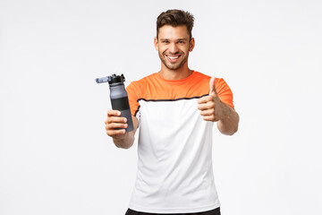 Fototapeta na wymiar Handsome mascular and healthy fitness instructor recommend drink more water as key to good shape. Smiling good-looking sexy sportsman in activewear show thumb-up and hold sports bottle