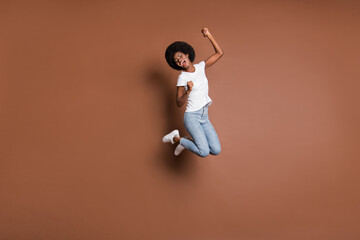 Fototapeta na wymiar Photo of energetic girl jump open mouth raise fists wear white t-shirt jeans shoes isolated brown color background