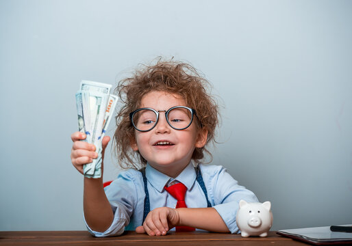 Cute Kid Girl Holding Dollars And Have An Idea How Earning Much Money In Crisis. Serious Child Teaching In Eyeglasses. Funny Happy Portrait Of Child Sitting At The Desk