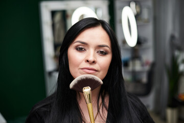 Close-up portrait of make-up artist at workplace in beauty studio, holding big make-up brush near face. Work process in beauty salon. Getting ready for gala dinner with help of professional stylist.