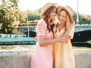 Two young beautiful smiling hipster woman in trendy summer sundress. Sexy carefree women posing on the street background in hats. Positive models having fun on embankment at sunset