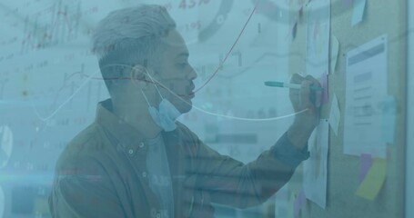 Animation of financial data processing over man with face mask writing on memo note - Powered by Adobe