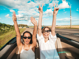 Portrait of two young beautiful and smiling hipster female in convertible car. Sexy carefree women driving cabriolet. Positive models riding and having fun in sunglasses outdoors.Enjoying summer days