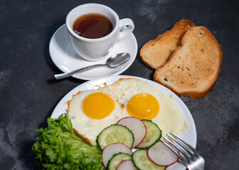Two fried eggs, lettuce, cucumber and radish slices on a white plate, toast and a cup of tea or coffee. Scrambled eggs. Breakfast. Healthy eating. Close-up
