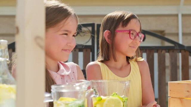 Waist-up shot of two happy mixed race girls of elementary age spending summer day outdoors selling lemonade at stand, giving high fives to each other