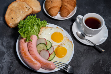 Two fried eggs and sausages, pieces of salad, cucumber and radish on a white plate, toast, croissants and a cup of tea or coffee. Scrambled eggs. Breakfast. Healthy eating. Close-up