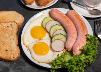 Two fried eggs and sausages, pieces of salad, cucumber and radish on a white plate, toast, croissants and a cup of tea or coffee. Scrambled eggs. Breakfast. Healthy eating. Close-up