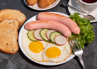 Two fried eggs and sausages, pieces of salad, cucumber and radish on a white plate, toast, croissants and a cup of tea or coffee. Scrambled eggs. Breakfast. Healthy eating. Close-up