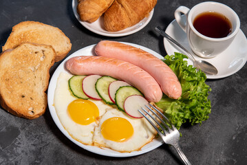 Two fried eggs and sausages, pieces of salad, cucumber and radish on a white plate, toast, croissants and a cup of tea or coffee. Scrambled eggs. Breakfast. Healthy eating. Close-up