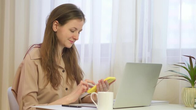 A woman in the office works at a laptop and reads the phone. using smart phone surfing social media, checking news, playing mobile games or texting messages. young smiles and looks into the phone. 