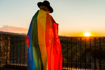 LGBT pride flag concept.Back view unrecognizable woman wrapped in rainbow gay flag at sunset.
