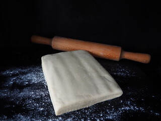 Raw puff pastry dough and wooden rolling pin on black background