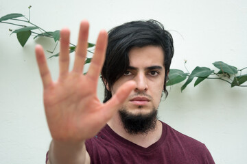 Portrait of a young caucasian black haired man with beard and red shirt looking at camera with his hand on the front
making a stop hand sign. White background and climbing plant with leaves behind
