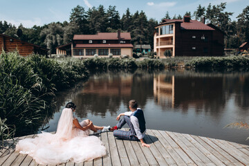 The newlyweds are resting on the wooden pier of a small lake. Hot summer day.