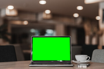 Mockup of laptop computer with empty screen with coffee cup and smartphone on table of the coffee shop background,Green screen