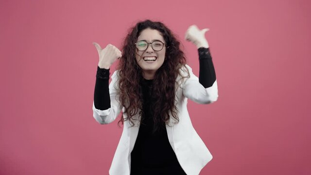 Happy young woman with curly hair, with her hands shows her thumb up as a sign, recommending smiling. Young hipster in white jacket and black shirt, with glasses posing isolated on pink background in