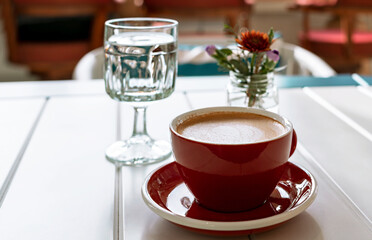 Coffee cappuccino in red cup, glass of water and small bouquet flowers in glass jar on white table