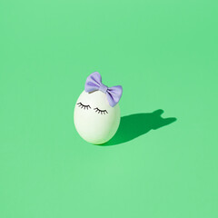 White Easter egg with eyelashes and purple bow tie headband on a light green background. Minimal creative holiday or food concept.
