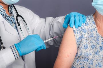 The doctor giving an injection to a senior woman wears a face mask. Elderly patient, being in a risk group, receiving COVID-19 vaccine in the hospital