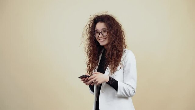 Happy young woman with curly hair holds a phone in her hand and vb with friends through the message. Young hipster in gray jacket and white shirt, with glasses posing isolated on beige background in
