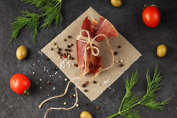 Pieces of spicy jerky tied with twine on parchment paper against a dark background. Herbs, spices and coarse salt, red cherry tomatoes, olives. homemade food