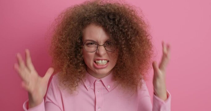 Outraged bothered woman shakes fists with rage stands furious complains about something has quarrel with someone wears spectacles and shirt isolated over pink background. Anrgry aggressive female
