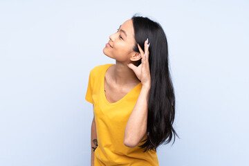 Teenager Chinese woman isolated on blue background listening to something by putting hand on the ear