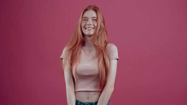 The young redhead with a beautiful smile, shows with her hand the gesture well, smiling happily at the room. 20-year-old freckled hipster teenager in a pink casual T-shirt, isolated on a pink