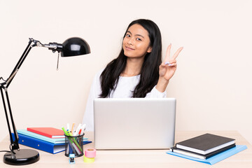 Student asian girl in a workplace with a laptop isolated on beige background smiling and showing victory sign