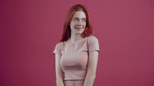 The shy young redhead, with her finger on her lips shows the sign to be silent, promises a secret. 20-year-old freckled hipster teenager in a pink casual T-shirt, isolated on a pink background.