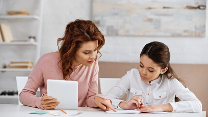 Woman with digital tablet pointing at notebook near daughter doing homework.