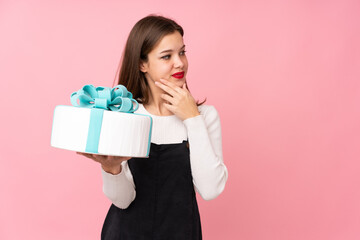 Girl holding a big cake isolated on pink background thinking an idea and looking side