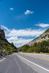 Paved road leading through canyon with green trees and bushes under a blue sky with white fluffy clouds.