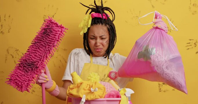 Unhappy housemaid looks with aversion at garbage bag and dirty mop poses near laundry basin busy doing housework wears rubber protective gloves has disgusting facial expression. Domestic chores
