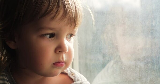 Portrait of a lonely upset toddler looking out the window. Orphan child sitting alone by the window.