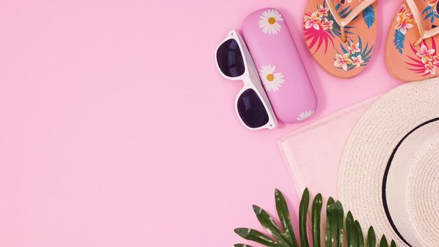 Summer beach accessories, hat, sunglasses and flip flops appear on pastel pink background. Stop motion flat lay