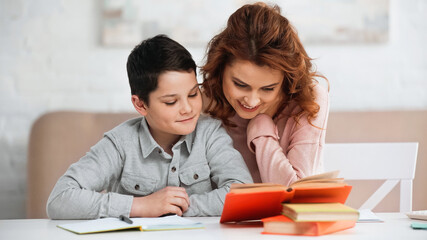 Fototapeta premium Smiling woman sitting near preteen son and books on blurred foreground.