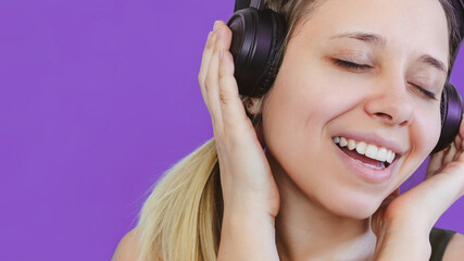 Close up of a young beautiful caucasian smiling happy cheerful blonde woman listening to music in large black headphones sings along on a purple background. Positive emotions and joy. Music fan