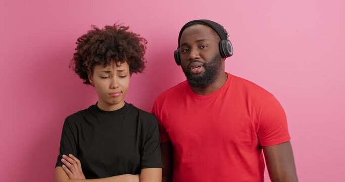 Relationship concept. Guilty man tries to speak with offended girlfriend asks for apologise wears stereo headphones on ears. Dissatisfied resentful Afro American woman crosses arms ignores husband
