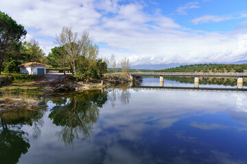 Fototapeta premium Landscape of a blue lake with trees and green plants, a little house on the shore and a bridge over the water. Atazar Madrid.