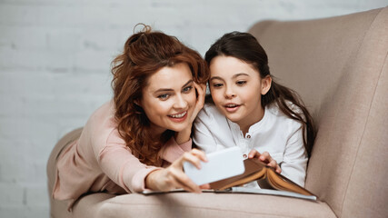 Smiling mother taking selfie with daughter near book on blurred foreground on couch.
