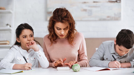 Pensive girl writing on notebook near mother with calculator and brother at home.