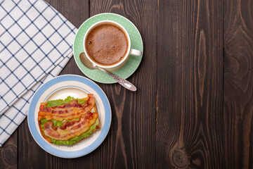 sandwich with fried bacon on a lettuce leaf and a cup of coffee top view