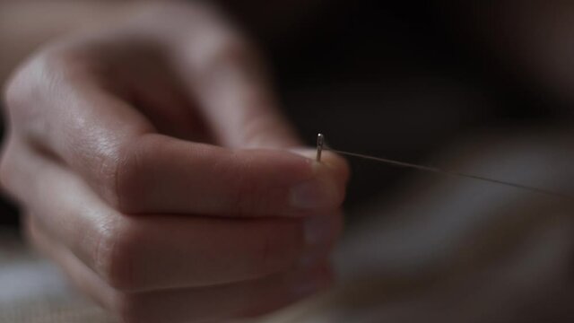 4K Close-up view of female hands inserting a thread into a needle hole for sewing. Young woman in green dress doing needlework. Hand made concept. Daylight.