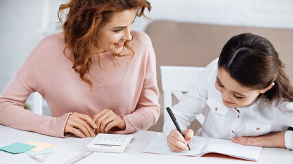 Smiling woman looking at girl writing on notebook during education at home.