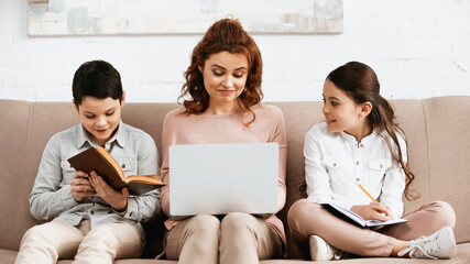 Smiling kids doing homework near mother with laptop on couch.