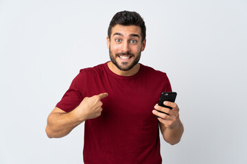 Young handsome man with beard using mobile phone isolated on white background with surprise facial...