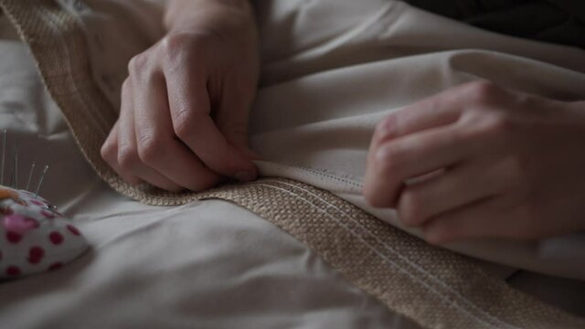 4K Close-up view of female hands rolling up beige fabric, takes pins from pin cushion and pin jute linen ribbon to cloth. Handmade concept. Boho rustic home decor. Young woman is sewing tablecloth.