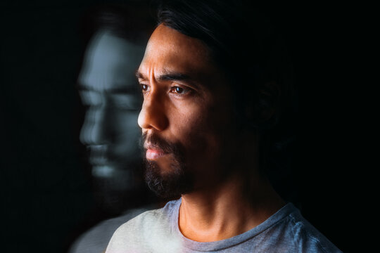 Close-up of the face of a Latin man and next to him his own depressed portrait with closed eyes. Long exposure image on black background. Depression concept
